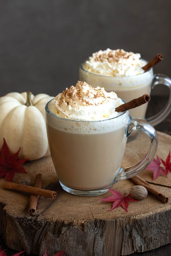 Two pumpkin lattes topped with whipped cream and cinnamon, beside a white pumpkin and autumn leaves.